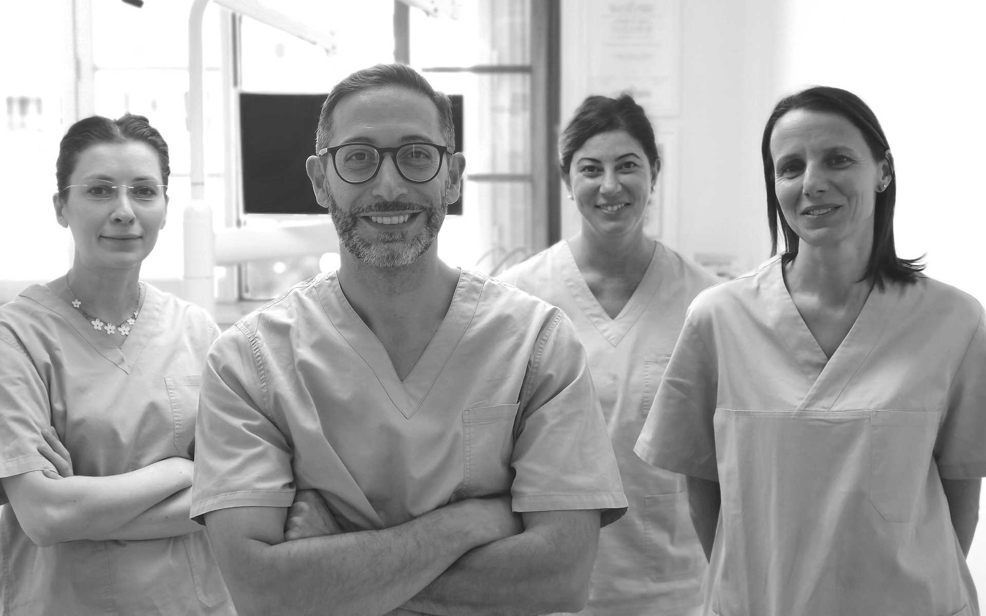 inner Quattro persone in camice stanno insieme in uno studio medico o dentistico, sorridendo e guardando la telecamera. L'immagine è in bianco e nero.
