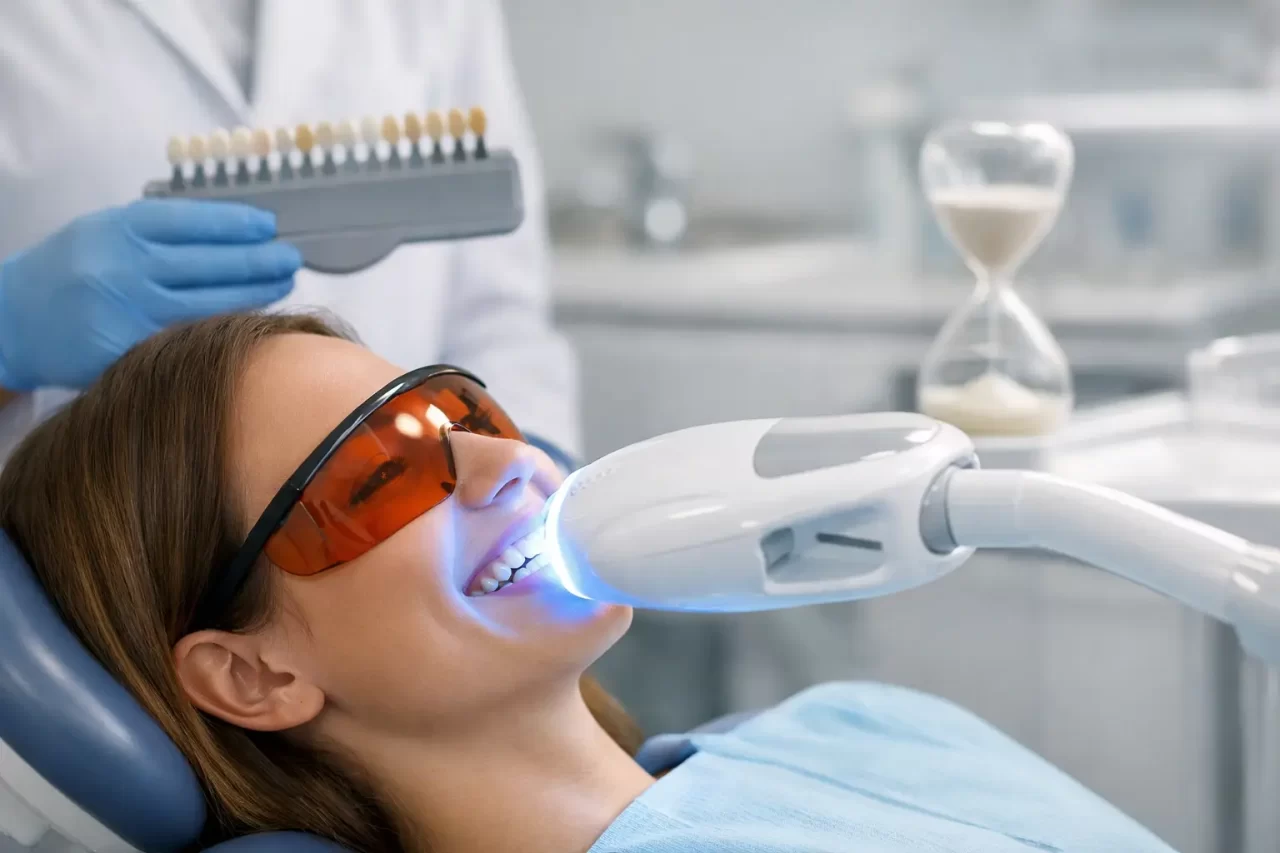 Una donna che indossa occhiali protettivi riceve un trattamento di sbiancamento dei denti in una clinica dentale. Un dentista tiene in mano una guida alla tonalità dei denti e una luce intensa illumina i denti della donna. Sullo sfondo è visibile una clessidra.