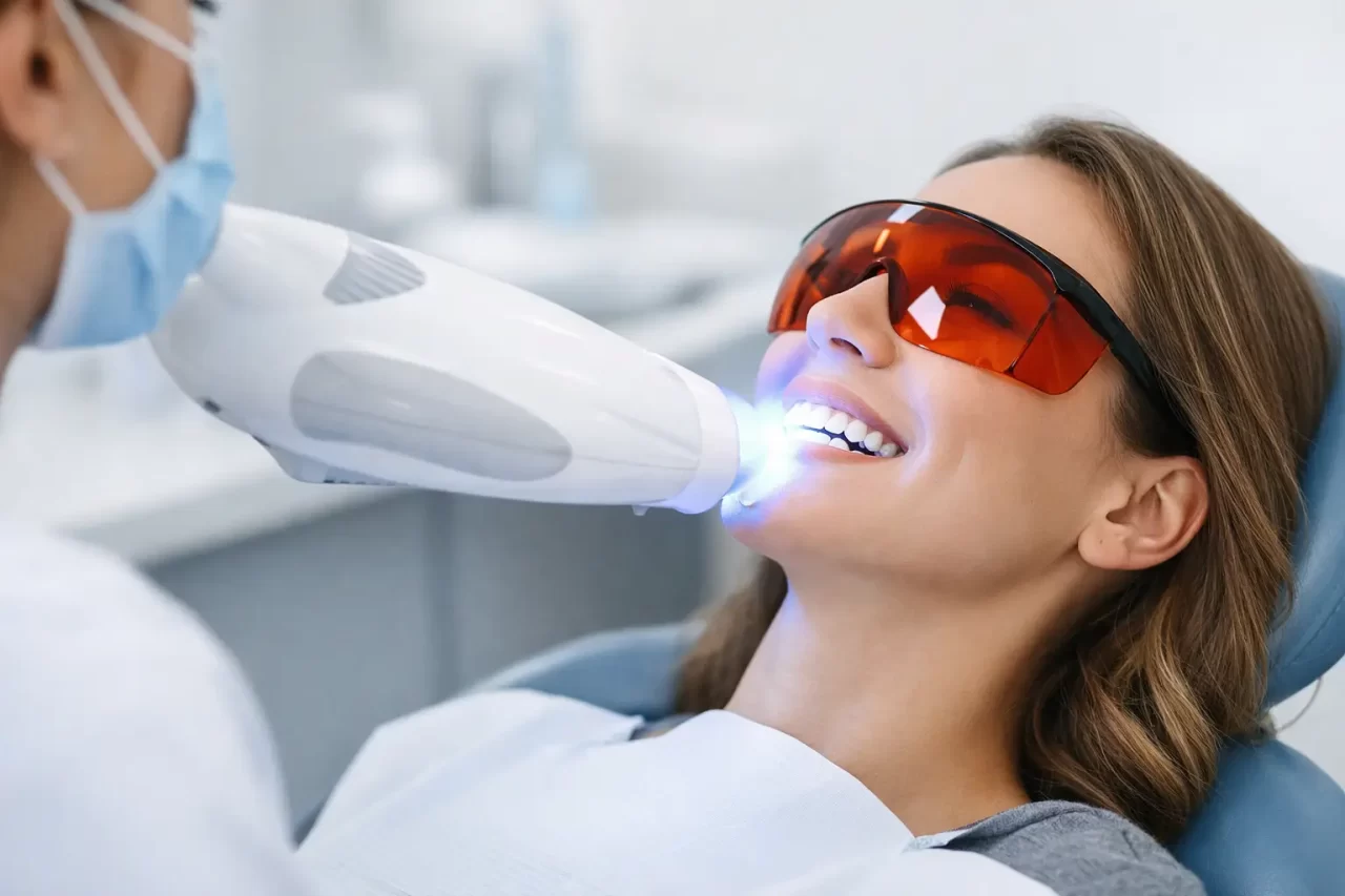 Una donna che indossa occhiali rossi protettivi riceve un trattamento professionale di sbiancamento dei denti con uno speciale dispositivo luminoso in una clinica dentale. Sorride ed è sdraiata sulla poltrona del dentista.